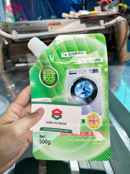 Túi có vòi in nền xanh lá hình máy giặt đựng nước giặt 500 g, cầm trên tay cạnh danh thiếp Đông Vũ